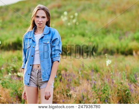 Happy Smiling Young Girl Wearing Blue Jeans Jacket And Shorts Is Standing In The Field