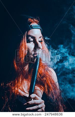 Red-haired Sexy Girl In A Cafe Dressed In Swimsuit And Cap Smoking A Hookah. Toned Image. Concept Of