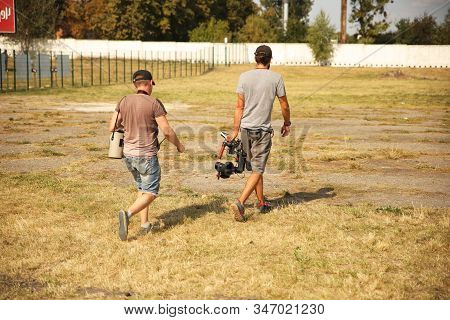 Bloggers Videographer And Photographer Go With The Camera On The Stabilizer On A Fenced Field With G
