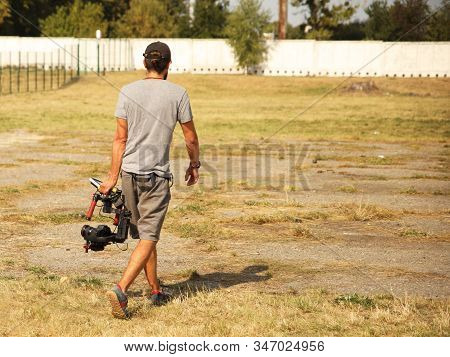 A Videographer Blogger Guy Is Walking With A Camera On A Stabilizer Over A Fenced Field With Grass. 