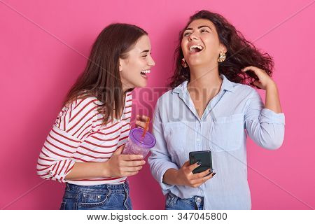 Horizontalshot Of Pretty Young Women Laughing Happily, Have Phone And Water Bottle In Hands, Wearing