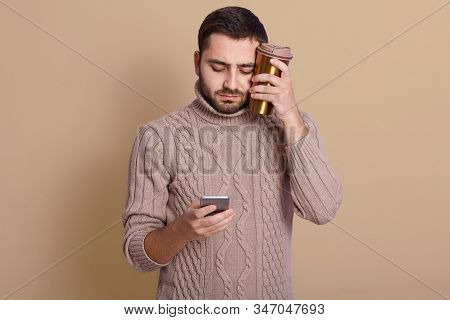 Indoor Shot Of Handsome Caucasian Man With Thermo Mug Of Hot Drink, Browsing Modern Smart Phone Agai