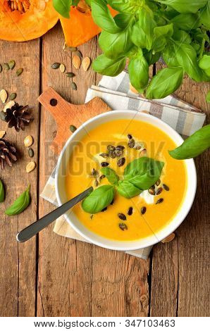 Bowl Of Cream Pumpkin Soup With Fresh Basil, Pumpkin Seed, Knife And Laddle On Rustic Wooden Table. 