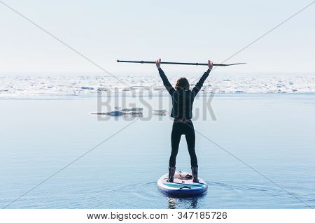 Slim Woman In Wetsuit Paddle On Sup Board And Holds Oar Over Her Head. Female Floating On Stand Up P