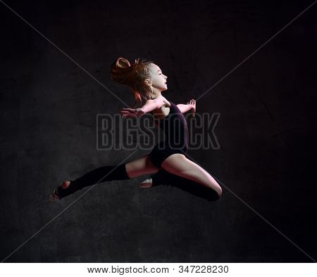 Young Girl Gymnast In Black Sport Body And Special Uppers Jumping In Gymnastic Pose Over Dark Backgr