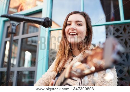 Portrait Of Young Female Musician Busking Playing Acoustic Guitar And Singing Outdoors In Street
