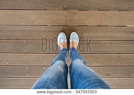 Female Legs In Denim Jeans Pants And Light Summer Sneakers Standing On Wooden Floow.