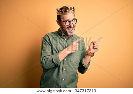 Middle age hoary man wearing king crown standing over isolated yellow background smiling and looking at the camera pointing with two hands and fingers to the side.
