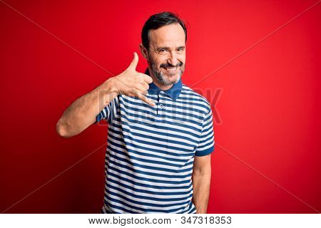 Middle age hoary man wearing casual striped polo standing over isolated red background smiling doing phone gesture with hand and fingers like talking on the telephone. Communicating concepts.