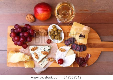 Assortment Of Cheese With Fruits, Grapes, Nuts, Glass With Wine And Cheese Knife On A Wooden Serving