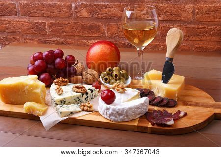 Assortment Of Cheese With Fruits, Grapes, Nuts, Glass With Wine And Cheese Knife On A Wooden Serving