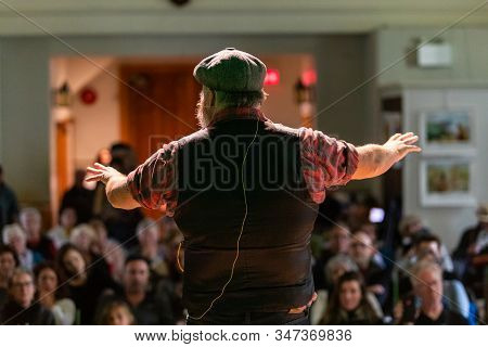 Rear View Of Male Speaker Wearing French Beret Cap And Giving Speech With Hand Gesture To The Audien