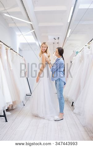 Smiling brunette wedding dress tailor assisting bride to wear dress between rows of beautiful gowns