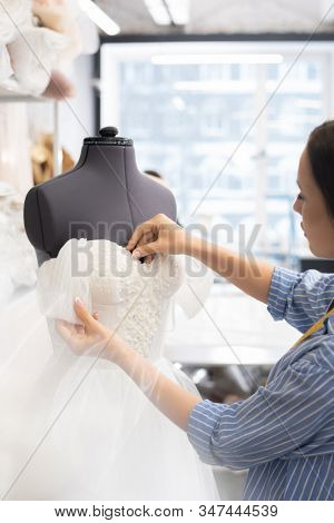 Serious young woman in casual shirt working on wedding dress corset in tailoring studio