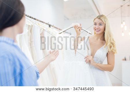 Excited beautiful bride with curly hair showing wedding dress to friend in shop