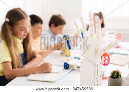Modern Technologies. Children Learning In Class With Humanoid Robotic Hand On Foreground, Panorama