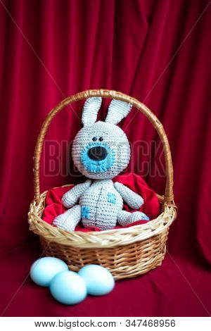 Knitted Gray And Blue Toy Cute Easter Bunny In Willow Basket And Blue Dyed Eggs On Red Textile Backg