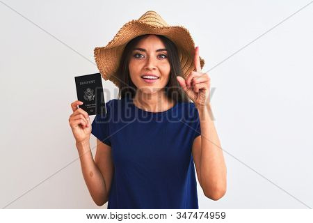 Beautiful tourist woman wearing hat holding USA passport over isolated white background surprised with an idea or question pointing finger with happy face, number one