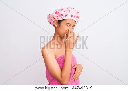 Young chinese woman wearing shower towel and cap bath over isolated white background smelling something stinky and disgusting, intolerable smell, holding breath with fingers on nose. Bad smells 