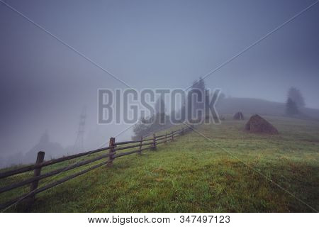 A Stunning View Of The Foggy Meadow In The Morning. Haycocks On The Grassy Field. Mysterious Pasture