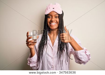 Young african american woman wearing pajama and eye mask drinking glass of water at night happy with big smile doing ok sign, thumb up with fingers, excellent sign