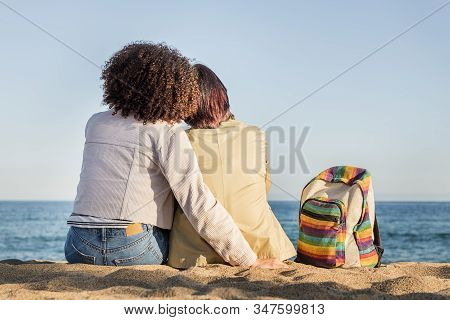Interracial Lesbian Couple Embracing In Front Of The Sea Next To A Backpack With The Rainbow Flag Co