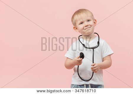 Child Blond Boy Stands And Holds A Stethoscope Or Phonendoscope In His Hands On A Pink Background Wi