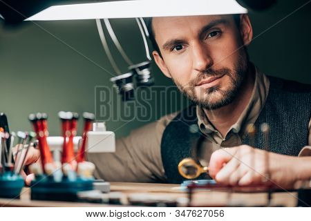 Handsome Watchmaker Looking At Camera While Working With Watch Parts And Magnifying Glass