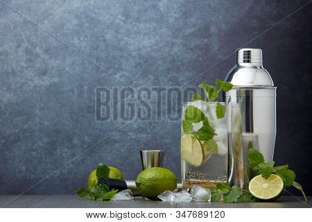 Set of bar tools: cocktail shaker, muddler, jigger, cocktail with ice in highball glass,  lime and mint on dark gray background with copy space. Bartender's tool. Mojito cocktail making.