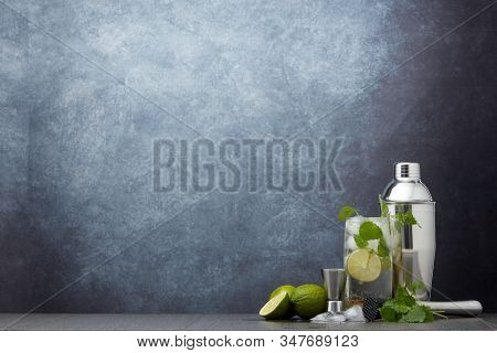 Set of bar tools: cocktail shaker, muddler, jigger, cocktail with ice in highball glass,  lime and mint on dark gray background with copy space. Bartender's tool. Mojito cocktail making.