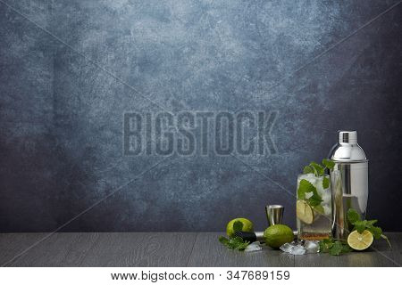 Set of bar tools: cocktail shaker, muddler, jigger, cocktail with ice in highball glass,  lime and mint on dark gray background with copy space. Bartender's tool. Mojito cocktail making.