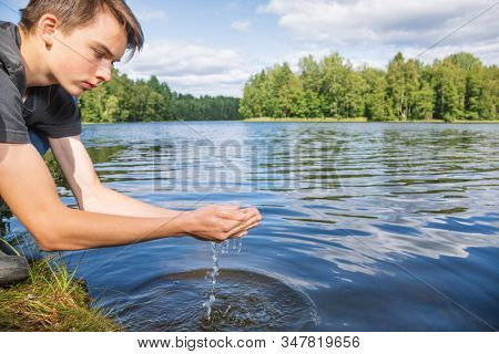 Teenage boy scoops up raw unfiltered water from a lake. Natural water source concept