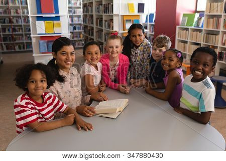 Front view of happy female teacher and schoolkids looking at camera while studying at table in school library