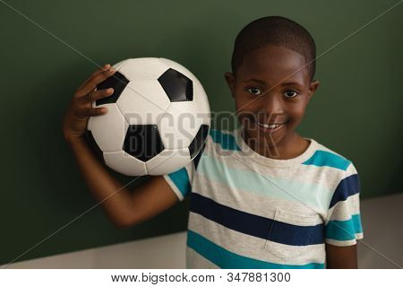 Front view of black schoolboy holding football and looking at camera in classroom of elementary school