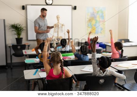 Rear view of school kids raising hand and sitting at desk in elementary school