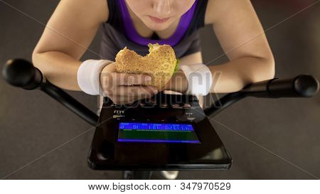 Corpulent Female Eating Fatty Burger While Riding Stationary Bike, Fast Food