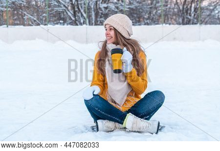 Portrait Of Happy Young Woman In Yellow Sweater And Ice Skates Sitting On The Snow, Holding Mug With