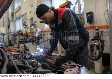 Contemporary automechanic in workwear bending over open car hood while chacking and repairing engine in garage