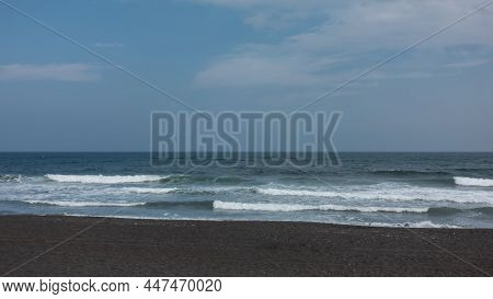 Long Waves Of Surf Roll Onto The Shore Of The Pacific Ocean And Foam. Black Volcanic Sand On The Bea