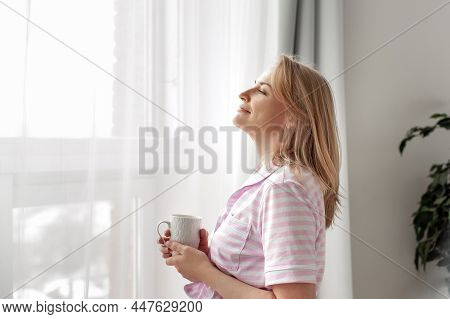 An Attractive Middle-aged Woman In Striped Pink Pajamas Stands By The Window With A Cup Of Hot Drink