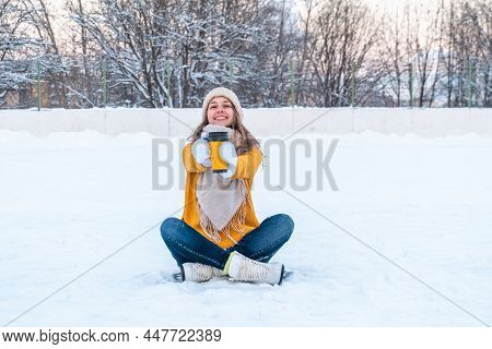 Portrait Of Happy Young Woman In Yellow Sweater And Ice Skates Sitting On The Snow And Holding Mug W