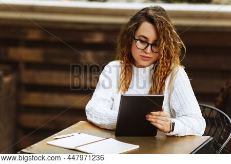 Young Female Student In Glasses Sitting At The Table, Using Tablet When Studying Or Working At Cafe,