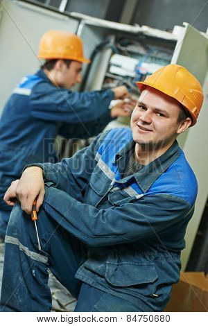 Happy young adult electrician builder engineer in front of his co-worker screwing equipment in fuseboard