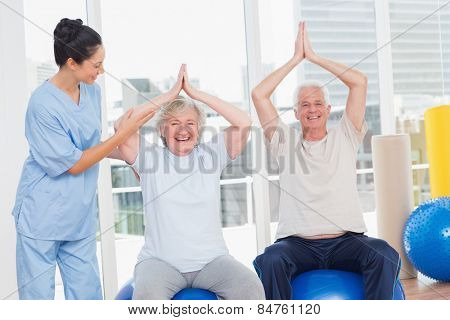 Portrait of senior couple on exercis ball being assisted by trainer in gym
