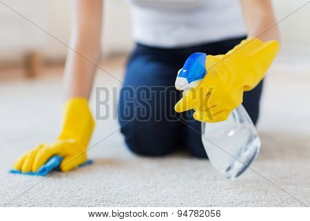 people, housework and housekeeping concept - close up of woman in rubber gloves with cloth and detergent spray cleaning carpet at home
