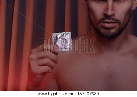 Cropped shot of young man with naked torso holding condom in bedroom. Safety sex concept.