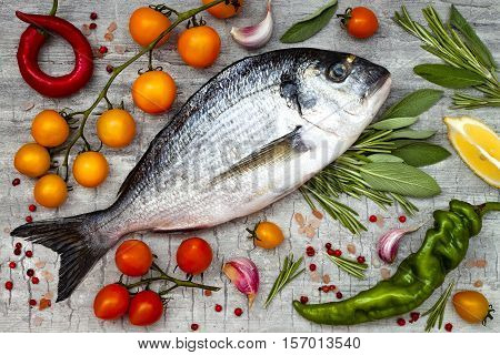 Fresh uncooked dorado or sea bream fish with lemon aromatic herbs vegetables and spices over grey stone background. Top view