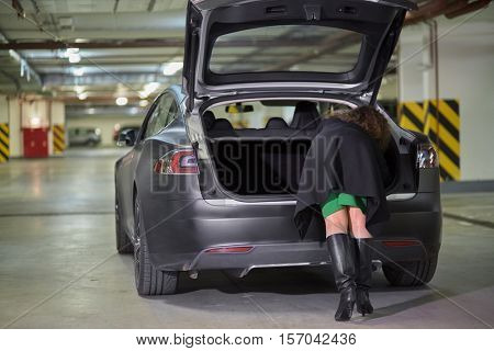 Young woman rummages in trunk of car standing at underground parking, rear view.