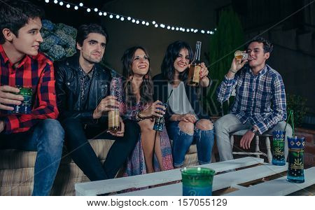 Happy young friends laughing and talking while woman showing beer at camera in a outdoors party. Friendship and celebrations concept.