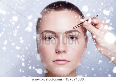 Young woman with short hair plucking eyebrows with tweezers close up woman plucking eyebrows depilating with tweezers. beauty people winter and health concept . over snow on gray background